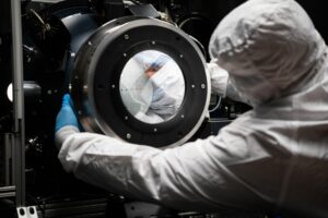 A man wearing a white clean room suit is seen from the shoulders up. He is facing away from the camera, arms outstretched and holding onto a large piece of a scientific instrument. The instrument is circular shaped. There is a circle in the center that is surrounded by another ring of a black circle. The center circle reflects the face of the man, who has a mask that covers his nose and mouth.