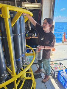 A woman stands in the image facing toward the left. She is wearing a dark brown shirt and teal colored pants. Her right arm is lifted up toward a piece of machinery that is made of several cylindrical tubes and is surrounded by bright yellow piping. 