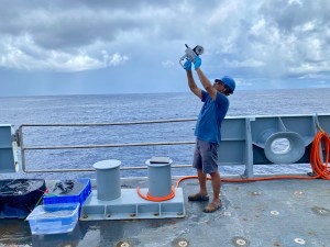 A man stands on board a ship facing the left side of the image. He is wearing a blue hard hat, a blue tshirt, gray shorts, and is holding a scientific instrument above his head, pointing it towards the sky. On the boat are tables and buckets next to the man. Just behind him are the railings of the side of the ship. The background of the image shows the flat blue water of the ocean, the horizon, which is about two thirds of the way up the image, and a gray-blue sky covered in clouds.