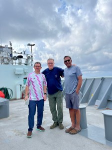 Three men stand on a boat, the man in the middle has his arms around the backs of the other two, all three facing the camera and smiling for the picture. The man on the left is wearing glasses, a white short sleeved shirt with a pink and pale green stripe pattern and blue jeans. The man in the middle is wearing glasses, a blue shirt and green pants. The man on the right is wearing sunglasses, a light blue shirt, and gray shorts. Behind them on the ship are railings to the edge of the ship as well as several pieces of machinery and equipment. The background of the image shows a blue-gray sky covered in clouds.