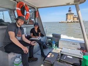 A woman and a man are sitting on a bench on a boat on the left side of the image. The man is sitting closer to the camera and is wearing a hat, a gray t-shirt, and black pants. The woman is wearing a hat, a black t-shirt, and pants. There is equipment and boxes in front of them, and an orange, circular life preserver hanging behind them. In the background are the brown-blue waters of the lake, as well as a brown building sitting on an island on the lake.