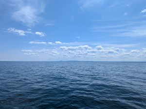 The image is split by the horizon down the middle. The bottom part of the image is of the dark blue waters of the lake. There are slight ripples on the water, but no waves. The top portion of the image shows bright blue skies with some wispy clouds, primarily closer to the horizon. 