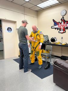 Two men stand centered in the image, facing each other. The man to the left, wearing jeans, a t-shirt, and a hat, helps secure a bright yellow spacesuit onto the man to the right. 