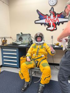 A man wearing a yellow spacesuit sits in a chair, his left arm outstretched reaching for a glove that another man just at the edge of the image is holding for him. The man in the chair also has a space helmet on and is smiling at the camera. 