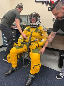 A man in a yellow spacesuit sits in a chair, his eyes closed, and the spacesuit pressurized to look fully inflated. Two other men stand to his left and right checking seals on the spacesuit.