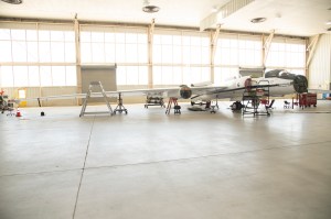 A plane sits in a hangar. The front nose of the plane is removed. The body of the plane is seen on the right side of the image and one of the wings extends out to the left part of the image. The background shows several windows allowing a bright light into the room.