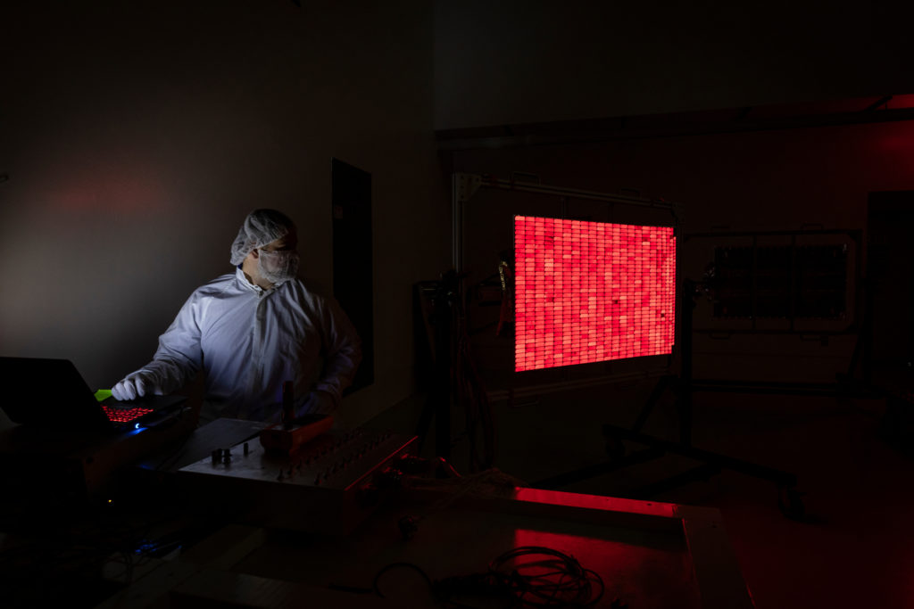 A person wearing a clean suit sits in a dark room looking a solar panel, on which all the cells are glowing red.