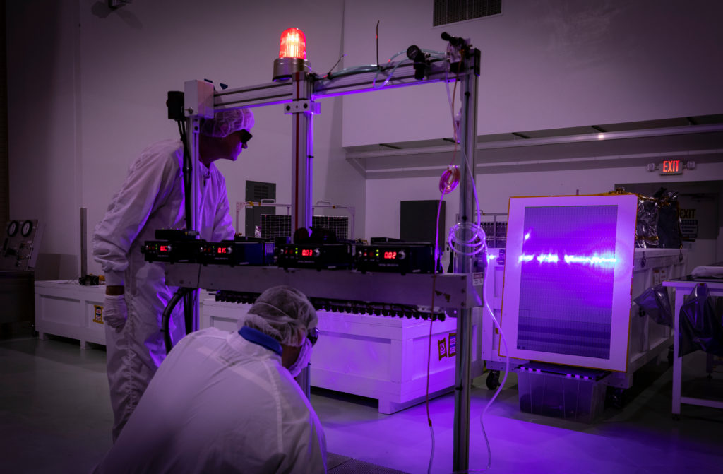 Two people in clean suits shine a purple laser on a solar panel.