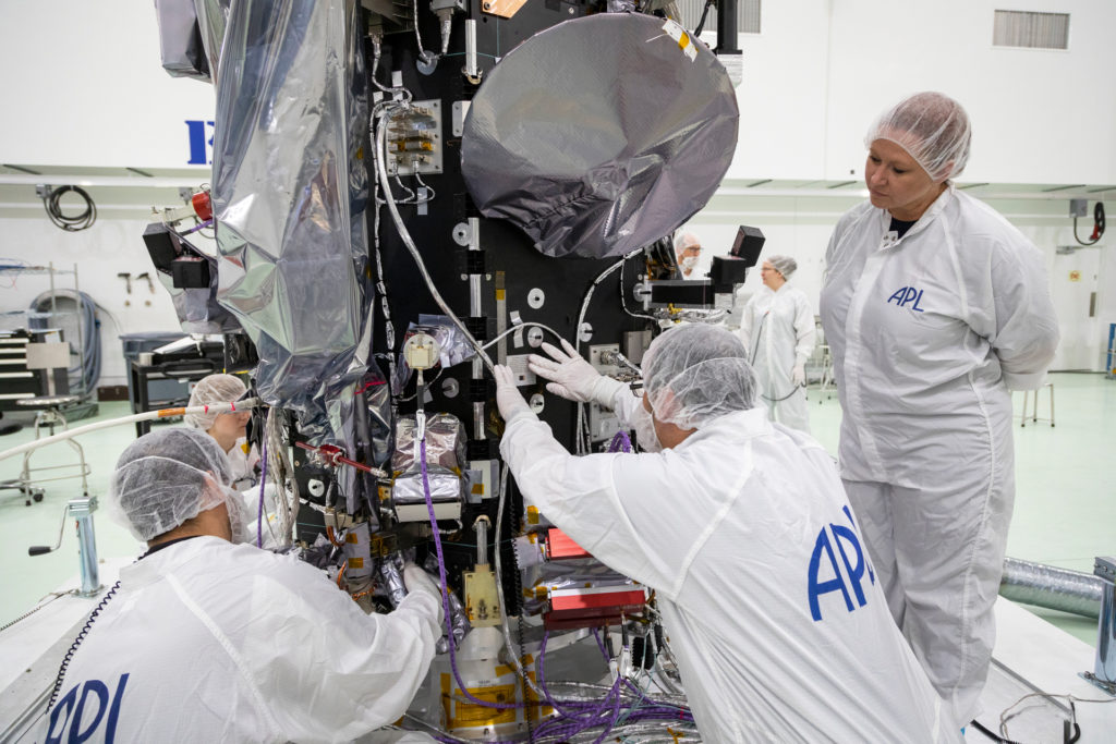 A person in a clean suit places a plaque on the side of the Parker Solar Probe spacecraft, while three other clean-suited people look on. 