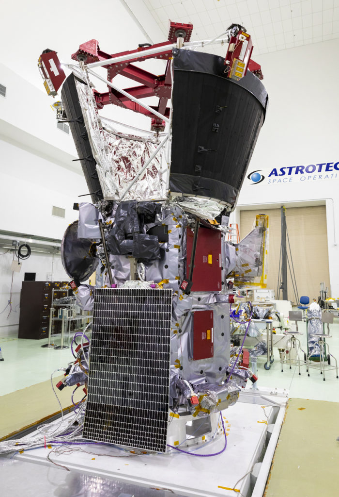 A spacecraft sits in a clean room with a solar array installed on the side.