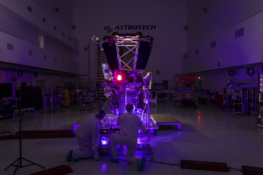 Two people in bunny suits kneel in front of a solar panel attached to a spacecraft while operating a purple laser in the dark.