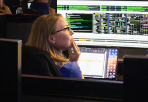 A woman sits in front of a laptop and a computer screen, looking into the distance, with her hand on her chin.