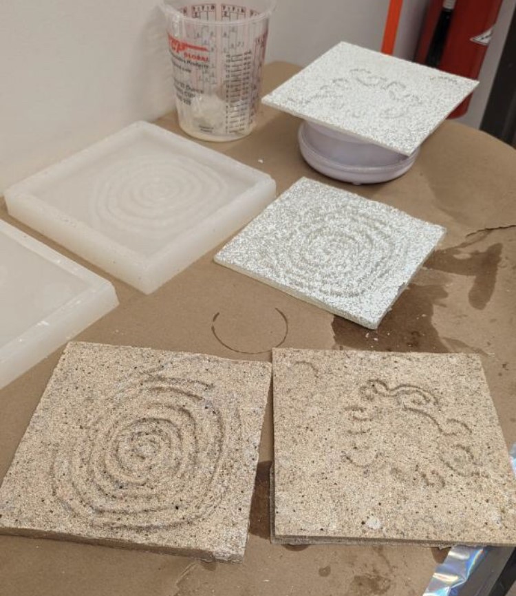 Two light-brown square presentations of petroglyphs sit on a brown table on the bottom of the image. To the left are two off-white rubber molds for the tactile representations. At the top of the image are two white, square petroglyph representations.