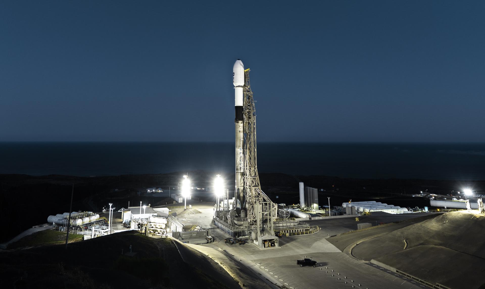 A SpaceX Falcon 9 rocket sits on a launch pad at dawn. Lights illuminate the rocket from below and the sky is just beginning to lighten on the horizon.