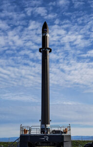 A full view of a black Electron rocket standing vertical on the launch pad framed by blue sky and wispy clouds. Photo: Rocket Lab