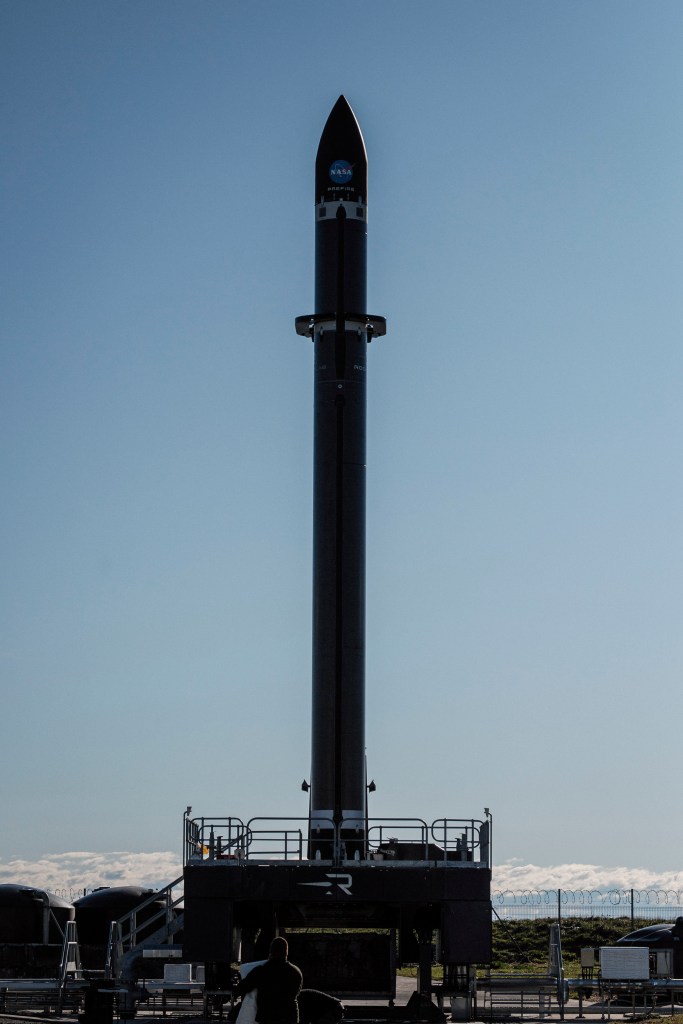 A Rocket Lab Electron rocket is erect on the launch pad on the coast of Mahia, New Zealand. The sky is blue and the sea is calm on the horizon behind the pad.