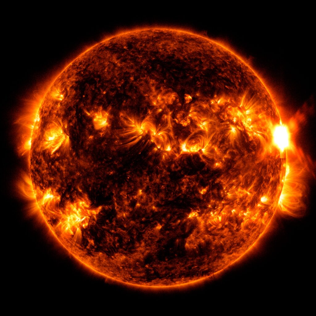 Il Sole mostrato in una tonalità rosso-arancio, con regioni inattive più scure e regioni attive luminose in arancione brillante.  Sul lato destro del Sole c'è un brillante lampo di luce: il bagliore solare.