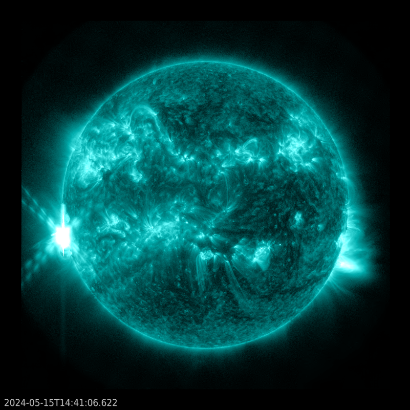 The Sun, shown in teal, appears against a black background. Swirling across the Sun are many bright teal active regions. On the left, a bright flash of teal and white light, forming an "X" shape, can be seen on the edge of the Sun against the black background.