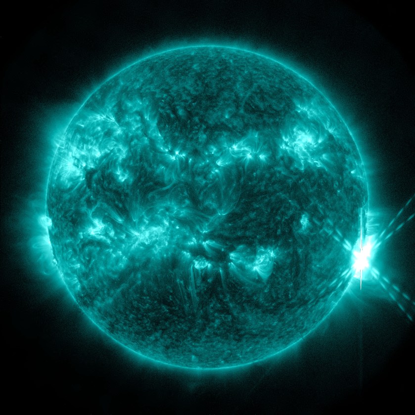 The Sun, shown in teal, against a black background. Swirling across the Sun are many bright teal active regions. On the right, a bright flash of teal and white light, forming an "X" shape, can be seen against the Sun and the black background.