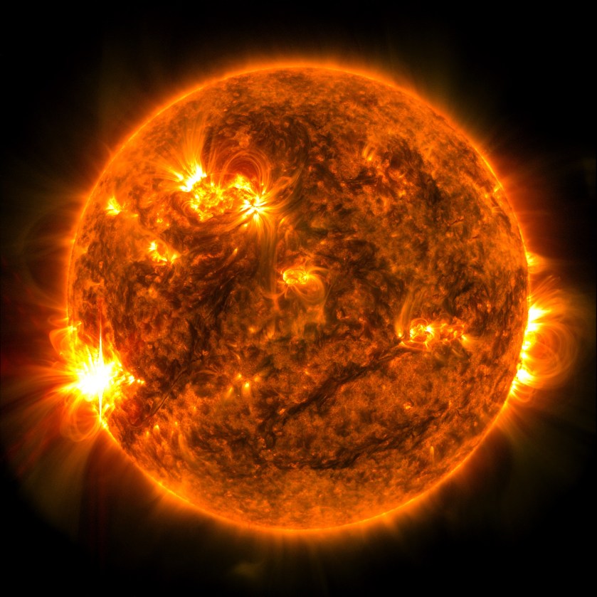 The Sun, shown in orange, appears against a black background. A few bright yellow active regions appear across the Sun. A bright flash of yellow and white light can be seen on the lower left edge of the Sun against the black background. 