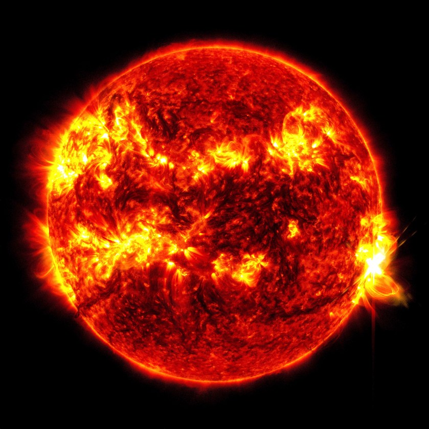 The red and yellow Sun against a black background. The Sun is primarily red with several bright yellow active regions swirling across its surface. On the right, a bright yellow burst is seen cascading off the Sun against the black background.