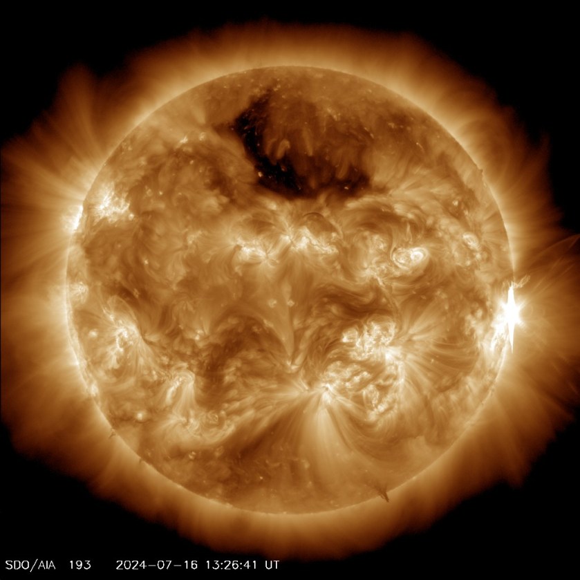 The Sun, shown in gold, against a black background. On the right is a bright white flash, on the limb of the star. The Sun has swirls of bright gold and darker gold regions, and one very large, dark black splotch toward the top.