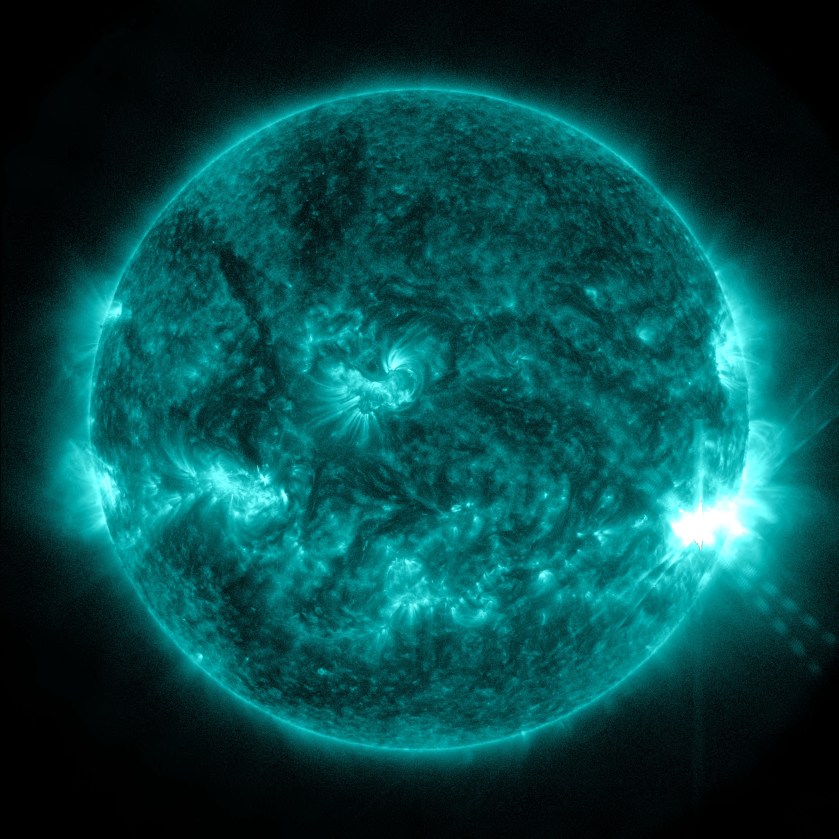 The Sun appears in shades of teal with some brighter and darker regions, set against a black background. On the far right side of the Sun is a bright flash of white, a solar flare.