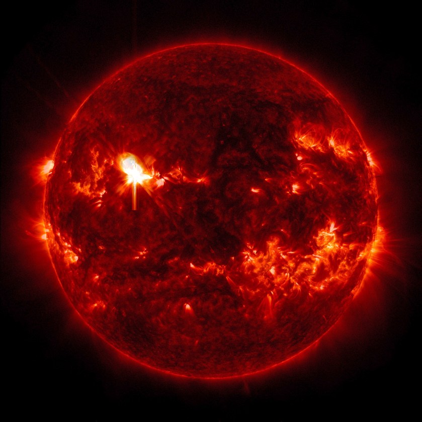 The Sun appears in shades of red with some brighter and darker regions, set against a black background. In the upper left part of the Sun is a bright flash of white, a solar flare.