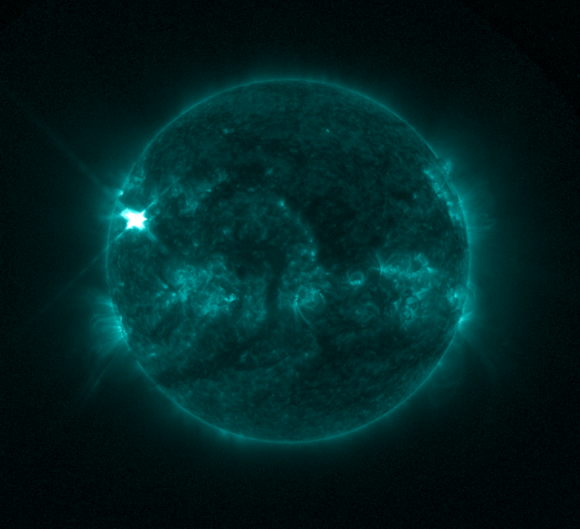 The Sun appears in shades of green with some brighter and darker regions, set against a black background. In the upper left part of the Sun is a bright flash of white, a solar flare.