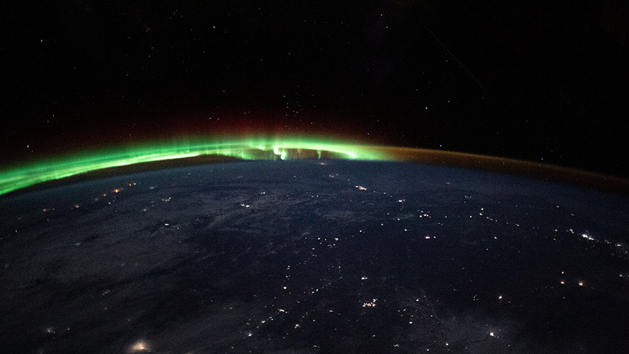 An aurora dances in the horizon of Earth's atmosphere as city lights shine through clouds cast over Mongolia.