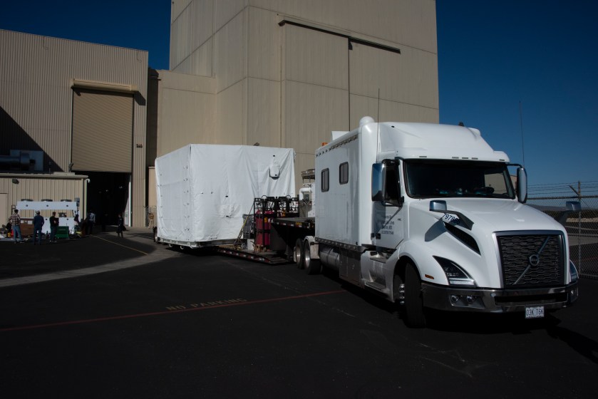 White truck with a white box on a trailer prepares to back into a tan hangar. The white box contains NASA's SPHEREx Spacecraft.