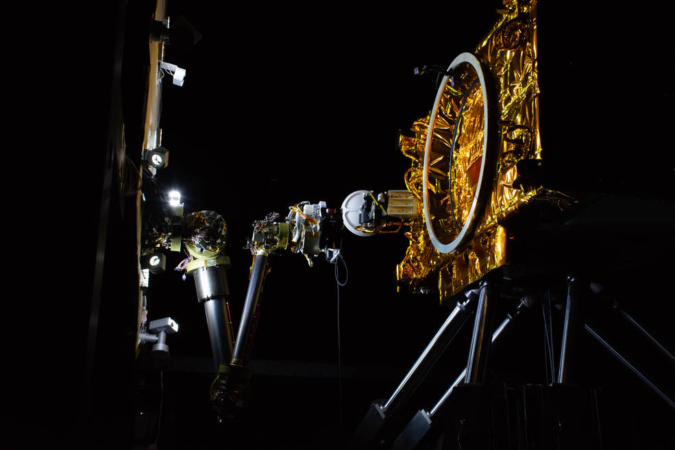 A six-legged, 10 by 16-foot robot mimics how satellites move in space. Sitting on top of the hexapod is a partial mockup of a satellite. Mounted to a panel close by, representing the payload deck of a robotic servicing spacecraft, is an advanced robotic arm.