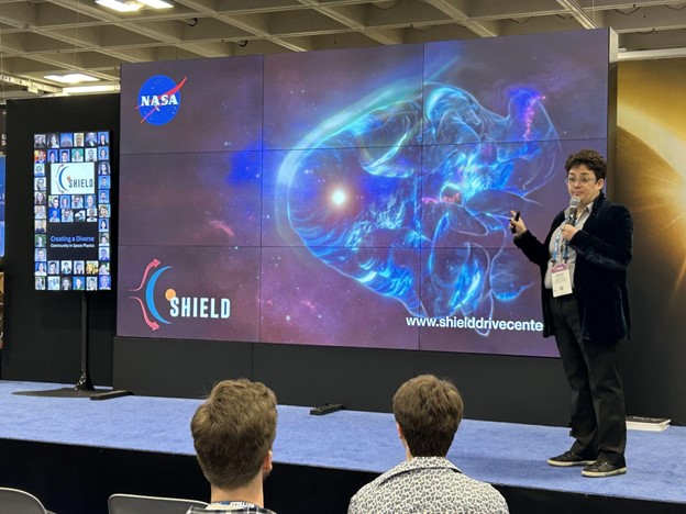 A woman in black clothing and a light grey shirt stands in front of a screen displaying a blue and grey artist's representation of what scientists believe the heliosphere looks like. 