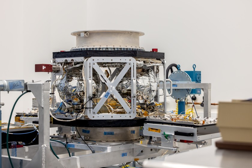 A white and black spacecraft, with a steel gray structure, sits on a steel gray stand in a white clean room.