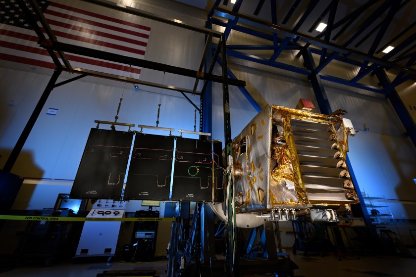 A silver and gold spacecraft with one solar array outstretched sits backlit in a large, dark white room.