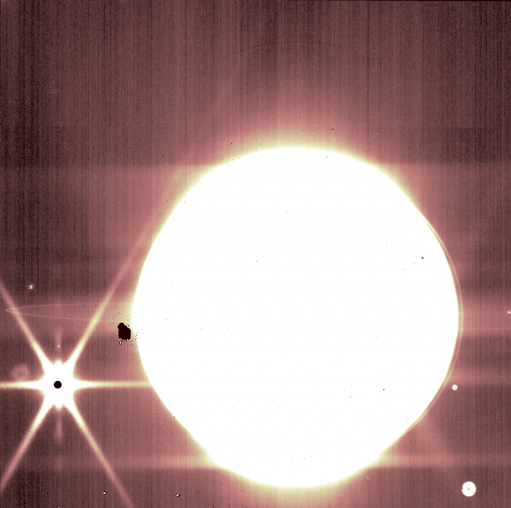 Jupiter is a bright white circle against a darker brown background. Moons are seen as small, white spots. Europa, to Jupiter's 8 o'clock, is a small black dot surrounded by bright white, with six white diffraction spikes.