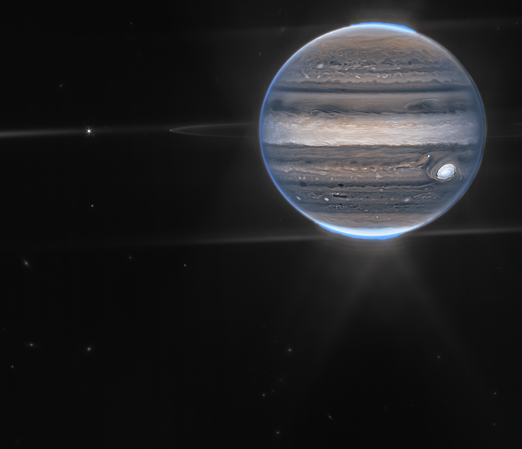 A wide-field view showcases Jupiter in the upper right quadrant. The planet’s swirling horizontal stripes are rendered in blues, browns, and cream. Electric blue auroras glow above Jupiter’s north and south poles. A white glow emanates out from the auroras. Along the planet’s equator, rings glow in a faint white. At the far left edge of the rings, a moon appears as a tiny white dot. Slightly further to the left, another moon glows with tiny white diffraction spikes. The rest of the image is the blackness of space, with faintly glowing white galaxies in the distance.