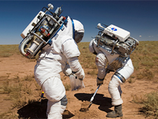 Two people dressed in spacesuits work in the desert